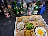 Photo showing various vintage empty soda bottles from brands including Atrea, 7up, Pepsi, and Ferro Bros Pottstown PA alongside multiple glass ashtrays with different shapes and colors.