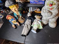 Five ceramic and porcelain dolls arranged on a dark surface, showing different styles and sizes including a clown figure and a Raggedy Ann type doll.