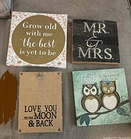 Photo of four decorative wall plaques: floral framed print with gold circle and inspiration quote, rustic 'MR. & MRS.' wood plaque, burlap hanging plaque with clip and moon quote, and green plaque with two owl figures and quote.