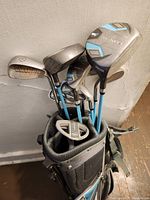 Full view of the golf club set including multiple clubs in a black golf bag leaning against a wall on a wooden floor.