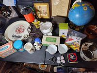Overview of various household items displayed on table including ceramic tray, globe, mugs, glass bowls, sewing accessories, and framed art.