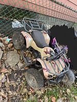 Four wheeler with beige, brown and red camouflage plastic body, black seat and black front and rear metal racks sitting outside on ground covered with leaves and rocks.