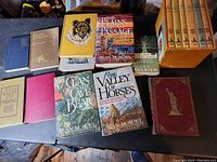 Photo showing 17 vintage books including Jean M. Auel novels (The Clan of the Cave Bear, The Valley of Horses, The Plains of Passage), Grace Miller White's From the Valley of the Missing, a small edition of The Hobbit by J.R.R. Tolkien, a set of 8 orange-spined books, and other miscellaneous titles with varied covers including a blue book and a green/gold book.