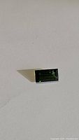 Close up photo of a bright green rectangular baguette cut tourmaline gemstone placed on a white surface, showing faceted edges and internal clarity.