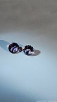 Close-up photo of two round amethyst gemstones with diamond cut facets, showing pink-purple colors under direct light on a neutral background.