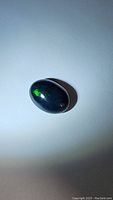 Oval cabochon jet black opal with slight green flash on white background, photo under natural or artificial light to show color play.