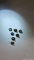Seven calibrated natural sapphires arranged on a light surface under a direct light source showing blue-green multicolored tones, oval cut, approximately 4x3mm each.