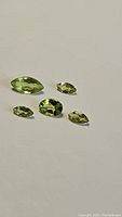 Photo showing five natural apple green peridot gemstones of mixed cuts arranged on a plain surface.