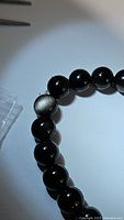 Close-up photo showing black polished round beads with one bead exhibiting a silver sheen natural flash characteristic of silver sheen obsidian.