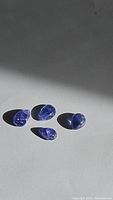 Four pear cabochon cut Tanzanite gemstones positioned on a grey surface showing their translucent purple blue color under natural sunlight.
