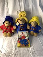 Three Paddington Bear plush toys and the Paddington at the Station novel arranged on a white cloth background.
