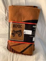 Folded brown fleece throw blanket with black printed design, wrapped with a label showing AC/DC and cannon logo.