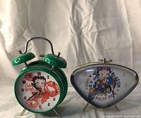 Two Betty Boop vintage style alarm clocks side by side on a white fabric background. Left green double bell clock with red illustration and right yellowish triangular clock with blue illustration. Visible crack on green clock protective glass.