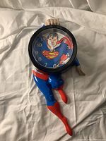 Clock showing full Superman figure holding clock face, displayed on white fabric background.