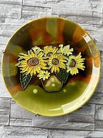 Front view of the circular ceramic plate showing the detailed hand painted sunflower design with green and orange glaze.