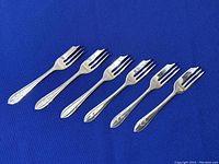 Set of six silver-plated dessert pastry forks arranged on blue fabric background showing front side and engraved patterned handles.