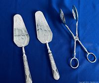 Three silver plated utensils on blue cloth: two cake paddles with floral engraving and one scissor style spoon spork.