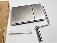 Cheese slicer with stainless steel surface, measurement grid and adjustable wire cutter, shown with original packaging and extra wire cutter in plastic bag.