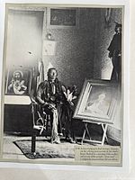 Front view of black and white photo showing Comanche Chief Quannah sitting in a room with two framed paintings, one religious image and one portrait, as well as a decorated interior.