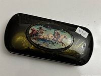 Top view of black lacquer case showing hand-painted oval scene of horse riders, with gold feather-like decorations around the edges.