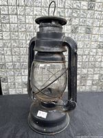 Photo of a vintage black metal oil lamp with a clear glass reservoir, metal handle, and visible signs of wear and age.