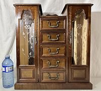 Front view of the vintage wooden jewellery box displaying the two glass-paneled side cabinets with floral etching and five central drawers with metal handles. Water bottle included for size reference.