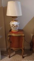 Side view of light wooden side table with cabinet door topped with decorative ceramic lamp with white shade.