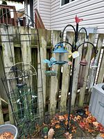 View of black metal wire plant holder with trellis, black metal multi-hook stand holding blue and brown bird feeders, and decorative elements.