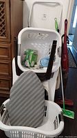 Photo showing 2 stacked white plastic laundry baskets, 2 white plastic chairs, a small ironing board inside one basket, green mop, red Hoover vacuum in background.