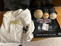 Photo showing white New York Yankees jacket folded, three baseballs with wear and markings placed on black fabric, one baseball has visible printed text.