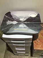 Photo showing plastic stackable storage containers with drawers, laptop holder with scenic mountain and lake print and cup holder, and a brown album titled 'Once Upon Album'.