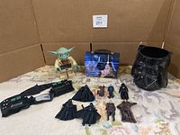 Overview of the lot showing a large Yoda figurine, 8 smaller Star Wars figurines, a blue Star Wars tin lunchbox, a black plastic Darth Vader helmet, and a Darth Vader belt with control panels.