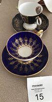 Two tea cup and saucer sets side by side showing black Royal Halsey set with nude ladies and cobalt blue Aynsley set with gold filigree.