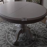 White round table with a central split and turned legs placed on a patterned rug in a room with a window and other furniture.