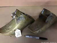 Front angled view of the pair of brass ceremonial stirrups showing embossed decorative patterns and attached tag with handwritten note.