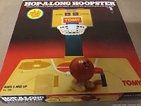 Front of box showing Tomy Hopalong Hoopster game with basketball character and scoreboard. Yellow plastic game court with hoop and wind-up basketball figure.