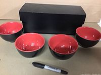 Four black bowls with red interior placed in front of a black box. A black Sharpie marker is placed for scale.