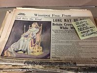 Photo showing pile of original Winnipeg Free Press newspapers featuring a large cover illustration of King George in regal attire and throne, with sticky note indicating coverage of King George and Queen Elizabeth.
