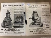Photo showing two original soda apparatus advertisements side-by-side with a pen for scale. The left ad is for the Arctic Soda Water Apparatus, showing a soda generator building and bottling outfit. The right ad is for the Torrid Soda Apparatus with ornate metal apparatus illustration.