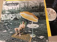 Photo of the Supertramp Crisis? What Crisis? vinyl record partially pulled from its album cover. The album cover depicts a man lounging under an umbrella with industrial ruins in the background. The vinyl record is black with a white label showing album details and RCA logo.