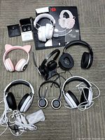 Overview of all headphones and earbuds in the lot arranged on floor, including boxed Redragon Zeus headset, black and white wired headsets, pink cat-ear headset, black headset with microphone, and three pairs of earbuds.