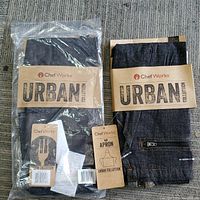 Two Chef Works Urban Collection aprons in original packaging and one unpackaged to show material and details.