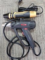 Two electric heat tools laid flat, one TGK heat gun, one black and gold device