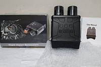 Night vision binoculars positioned upright next to the original box and user manual on white surface