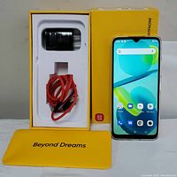 Smartphone lying on a table next to its yellow box containing charging adapter and red USB cable, along with a yellow phone case labeled 'Beyond Dreams'