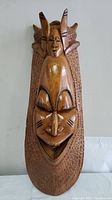 Front view of the wood carved Ibono tradition mask showing overall shape, facial features, and textured background.