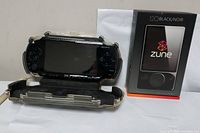Sony PSP portable game console displayed in clear hard shell case alongside Zune 120GB portable media player box and accessories on white background.