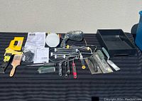 Wide photo showing the full set of painting equipment including Wagner Flexio 570 sprayer, paint rollers, brushes, trays, scrapers, and instruction manuals arranged on a black striped cloth.