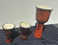 Three wooden hand drums of different sizes arranged against a gray background, showing entire drums and their natural skin heads and rope tuning.