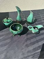Five pieces of green glazed pottery arranged on a dark cloth, showing vases, bowl, lidded jar, and candle holder.